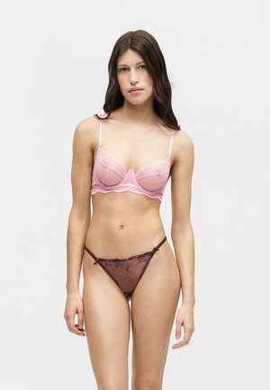 Frau mit langen braunen Haaren, die einen rosa Spitzen-BH und transparente dunkelviolette Spitzenunterwäsche trägt, steht vor einem schlichten hellen Hintergrund.