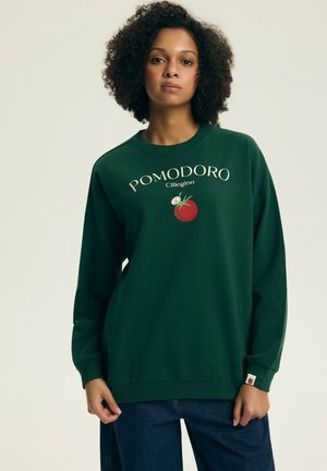 Kobieta o kręconych włosach, ubrana w ciemnozieloną bluzę z ilustracją pomidora i białym napisem "POMODORO Ciliegino".