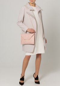 Light pink envelope handbag in textured material with a gold metal accent, paired with a light gray coat and a white dress.