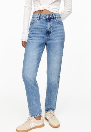 Kvinde iført lyseblå jeans med høj talje, beige sneakers og en hvid langærmet top bundet i en knude ved taljen.