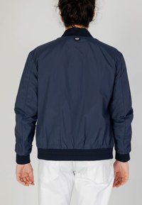 Giacca bomber blu navy con una texture liscia, polsini e orlo a coste, e un piccolo logo sul retro, indossata con pantaloni bianchi.