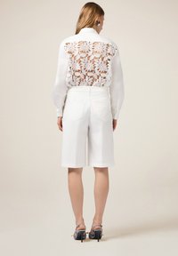 Blusa blanca con una espalda de encaje transparente que presenta patrones florales, combinada con pantalones cortos blancos. Colocada frente a un fondo neutro.