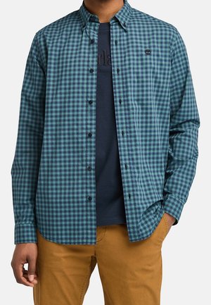Man wearing green and blue checkered button-up shirt over a dark navy graphic T-shirt and light brown pants, hand in pocket.