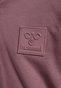 Écusson carré en caoutchouc avec logo hummel sur fond de tissu mauve texturé.