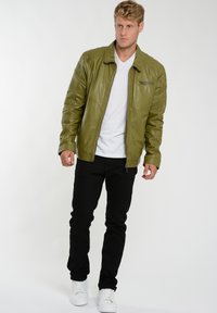 Olivgrüne Lederjacke mit einem Reißverschluss vorne, gerippten Schulterdetails und einer schlanken Form, kombiniert mit schwarzen Jeans und weißen Sneakers.