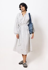 Gestreiftes, tailliertes Langarmkleid mit Knöpfen und einem Taillengürtel. Leichter Stoff, knielange Länge, kombiniert mit einer blauen Häkelhandtasche und gestreiften Schuhen.