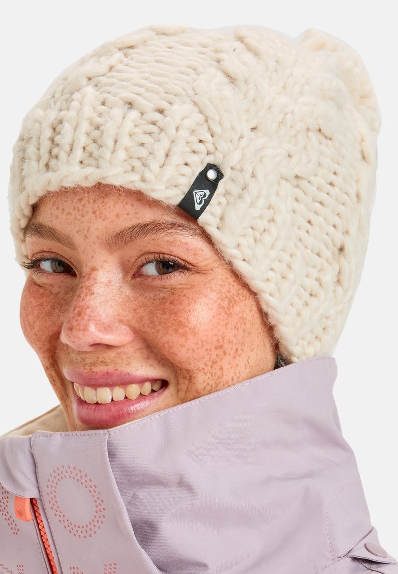 Gebreide crèmekleurige beanie met een dikke textuur en een klein zwart label aan de zijkant. De drager draagt een lichtpaarse jas met een opstaande kraag.