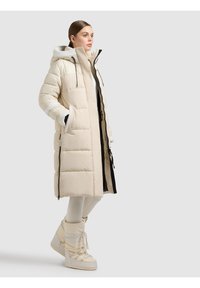 Lång beige vadderad jacka med huva, utrustad med svarta dragkedjedetaljer, sidofickor och ett mjukt foder. Matchas med beige knähöga stövlar.