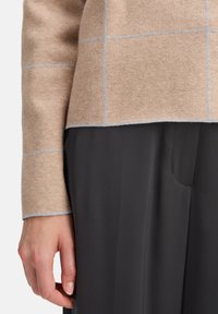 Die Hand einer Frau neben einem beigen Pullover mit hellen Karolinien und schwarzen Hosen, die die Textur und Passform der Kleidung hervorhebt.