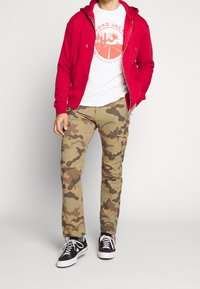 Sudadera roja con cremallera, camiseta gráfica blanca y pantalones cargo de camuflaje con bolsillos laterales, combinados con zapatillas negras con suelas y cordones blancos.