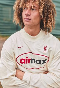 Maglia a maniche lunghe crema con colletto a costine. Presenta un grande logo "airmax" rosso e uno stemma LFC rosso sul lato sinistro superiore. Capelli ricci in vista.