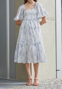 Biała i niebieska kwiatowa sukienka maxi z bufkami, kwadratowym dekoltem i warstwami falban. Noszona z pomarańczowymi sandałami na paski.