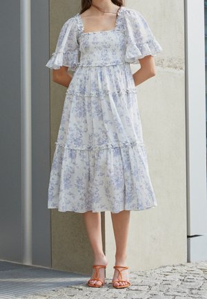 White and blue floral maxi dress with puff sleeves, a square neckline, and tiered ruffles. Worn with orange strappy sandals.