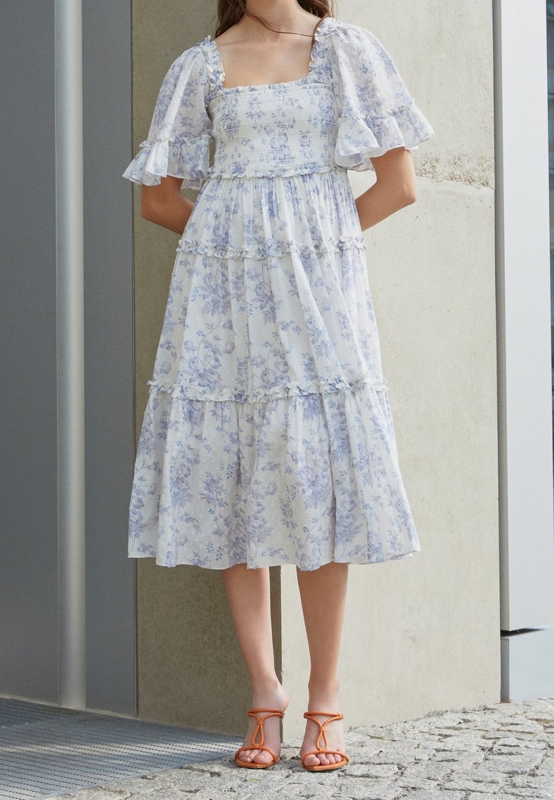 Biała i niebieska kwiatowa sukienka maxi z bufkami, kwadratowym dekoltem i warstwami falban. Noszona z pomarańczowymi sandałami na paski.