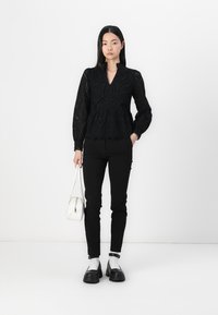 Black lace top with a peplum cut, features long sleeves, floral patterns, paired with black pants and chunky black shoes. Holds a white bag.