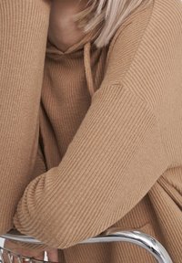 Hoodie beige a coste con una vestibilità rilassata. Presenta un cappuccio e un tessuto testurizzato. Braccia incrociate, appoggiato su una sedia in cromo.