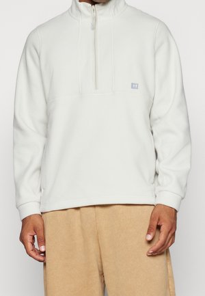 Hvid fleece half-zip pullover med opretstående krave, der har en brystsømm og et lille logo-patch på venstre side. Tekstureret stoffinish.