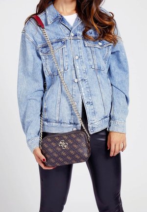 Hnědá kabelka přes rameno s metalickým řetízkovým popruhem, s logem na přední straně. Nohena s světle modrou džínovou bundou a černými kalhotami.