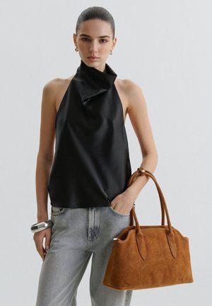 Donna che indossa un top nero senza maniche con collo all'americana e jeans grigio chiaro, che tiene in mano una borsa in camoscio marrone con due manici.