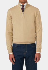 Maglione beige con zip e colletto, realizzato in tessuto leggero. Maniche lunghe e polsini a costine, abbinato a jeans in denim blu scuro.