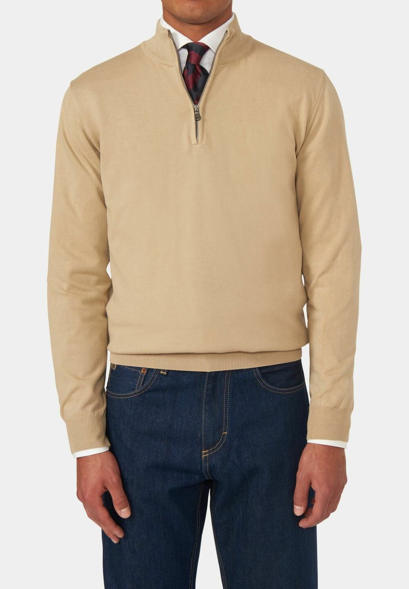 Maglione beige con zip e colletto, realizzato in tessuto leggero. Maniche lunghe e polsini a costine, abbinato a jeans in denim blu scuro.
