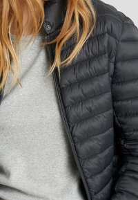 Sort quiltet pufferjakke med lynlåslukning, der har en høj krave og vandret syninger, båret over en lys grå sweatshirt.