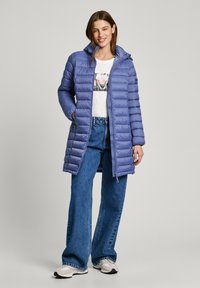 Padded purpurový kabát s vysokým límcem, zipovým uzávěrem a dvěma bočními kapsami. Nosený přes bílý tričko s grafickým potiskem a v kombinaci s modrými rozšířenými džíny.