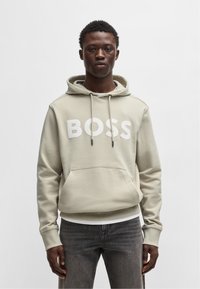 Šviesiai ruda mygtukinė palaidinė su dideliu baltu "BOSS" logotipu, su raišteliais reguliuojama kapisonu, priekyje yra kišenė ir raibės ant rankovių. Suderinta su tamsiomis džinsinėmis kelnėmis.