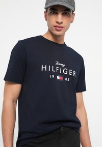 Mladý muž nosí šedou plochou čepici a tmavě modré tričko Tommy Hilfiger s logem a nápisem "1985" na přední straně.