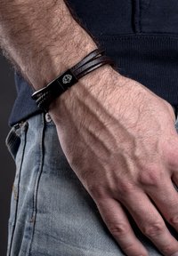 Black leather bracelet featuring multiple strands and a metal clasp with a lotus design. Worn on a hand against denim fabric.