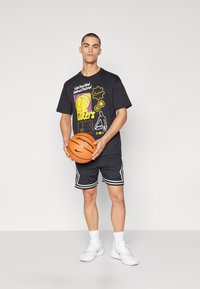 Černé grafické tričko s žlutým a fialovým potiskem, spárované s černými síťovanými šortkami s bílými akcenty. Drží oranžový basketbal. Bílé tenisky.