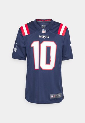 Person trägt dunkelblaues New England Patriots Football-Trikot mit der Nummer 10, hellgraue Jogginghose und schwarze Nike-Sneaker und steht vor weißem Hintergrund.