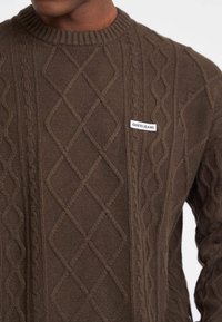 Brauner Kabelstrick-Pullover mit einem Rautenmuster, geripptem Ausschnitt und einem weißen Etikett mit der Aufschrift "GUESS JEANS" auf der linken Seite.
