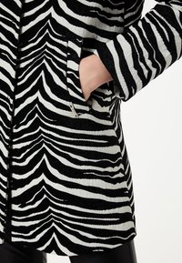 Mano en el bolsillo de un abrigo a rayas de cebra en blanco y negro con tela texturizada, llevado sobre leggings negros contra un fondo blanco.