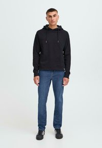 Zwarte hoodie met trekkoorden, gecombineerd met blauwe jeans. Model staat rechtop en draagt zwarte sneakers. Eenvoudig, casual ontwerp, stoftextuur zichtbaar.