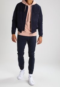 Giacca bomber navy con zip, felpa leggera rosa sottostante, jeans scuri, sneaker bianche. Design semplice, tessuto fluido, polsini e orlo elasticizzati.