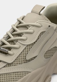 Beige sneaker featuring a mesh upper with smooth synthetic accents, flat laces, and a padded collar. Notable curved design elements throughout.