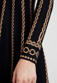 Manica di un abito in maglia nera con motivo a catena dorata, caratterizzata da dettagli intricati lungo il braccio e il polsino. Texture morbida.