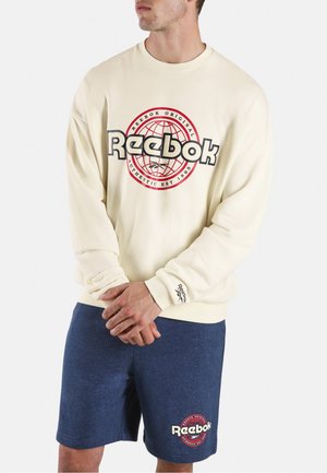 Mann trägt einen cremefarbenen Reebok-Sweatshirt mit rotem und schwarzem Globus-Logo und marineblaue Reebok-Shorts mit passendem Logo.