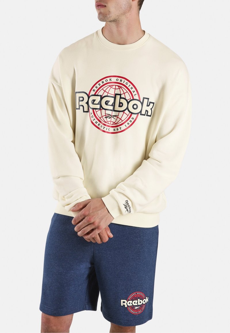 Man wearing a cream Reebok sweatshirt with a red and black globe logo and navy blue Reebok shorts with matching logo.