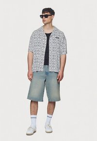 Άνδρας που φοράει μαύρα γυαλιά ηλίου, κοντομάνικο πουκάμισο με σχέδια πάνω από μαύρο μπλουζάκι, μακριές τζιν βερμούδες, λευκές κάλτσες και λευκά αθλητικά παπούτσια, όρθιος.