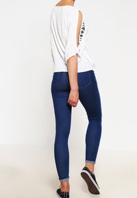 Top blanc avec des manches ouvertes et broderies noires, associé à un jean skinny bleu foncé, avec une taille mi-haute et des poignets retroussés.