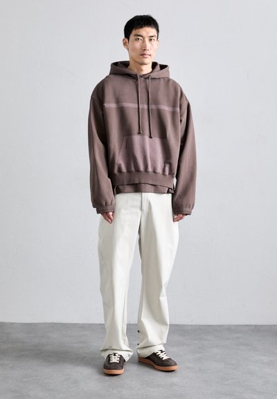 Sudadera con capucha marrón y un bolsillo frontal, con una franja horizontal, combinada con unos pantalones ligeros y holgados y zapatillas marrones.