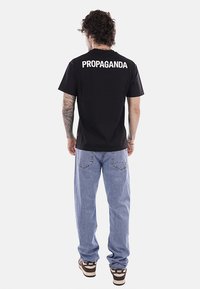 Uomo con capelli ricci in piedi di spalle, indossa una maglietta nera con la scritta "PROPAGANDA", jeans azzurri e sneakers Nike. Tatuaggi visibili sul braccio sinistro.