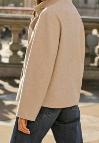 Beige wool-blend jacket with a structured design, large collar, and side pockets, paired with dark denim jeans. Soft texture, clean lines.