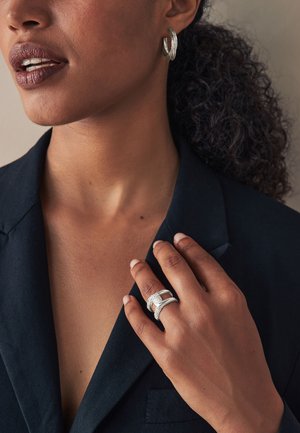 Woman with dark curly hair in a black blazer touching her chest, wearing textured silver hoop earrings and two silver rings on her finger.