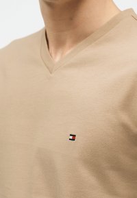 Μπεζ μπλούζα με V λαιμόκοψη σε ένα άτομο, με μικρό τετράγωνο λογότυπο σε κόκκινο, λευκό και ναυτικό μπλε χρώμα στο στήθος.