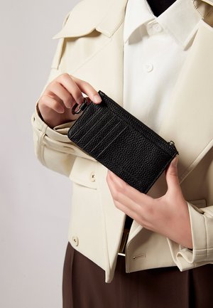 Sort tekstureret pung holdt i hånden, med lynlås og kortlommer på den ene side. Stående mod en lysfarvet jakke.