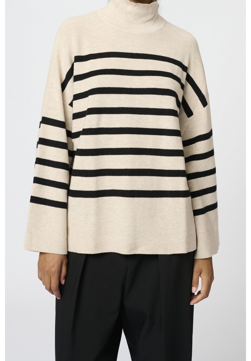 Osoba w beżowym swetrze z golfem w czarne poziome paski, w połączeniu z luźnymi czarnymi spodniami, na tle jednolitego jasnego tła.