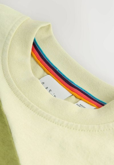 Nærbillede af en lysegul crewneck-skjortekrave med indvendig kant af flerfarvet (blå, lilla, pink, orange) stof og synlige vaskeanvisningstag.